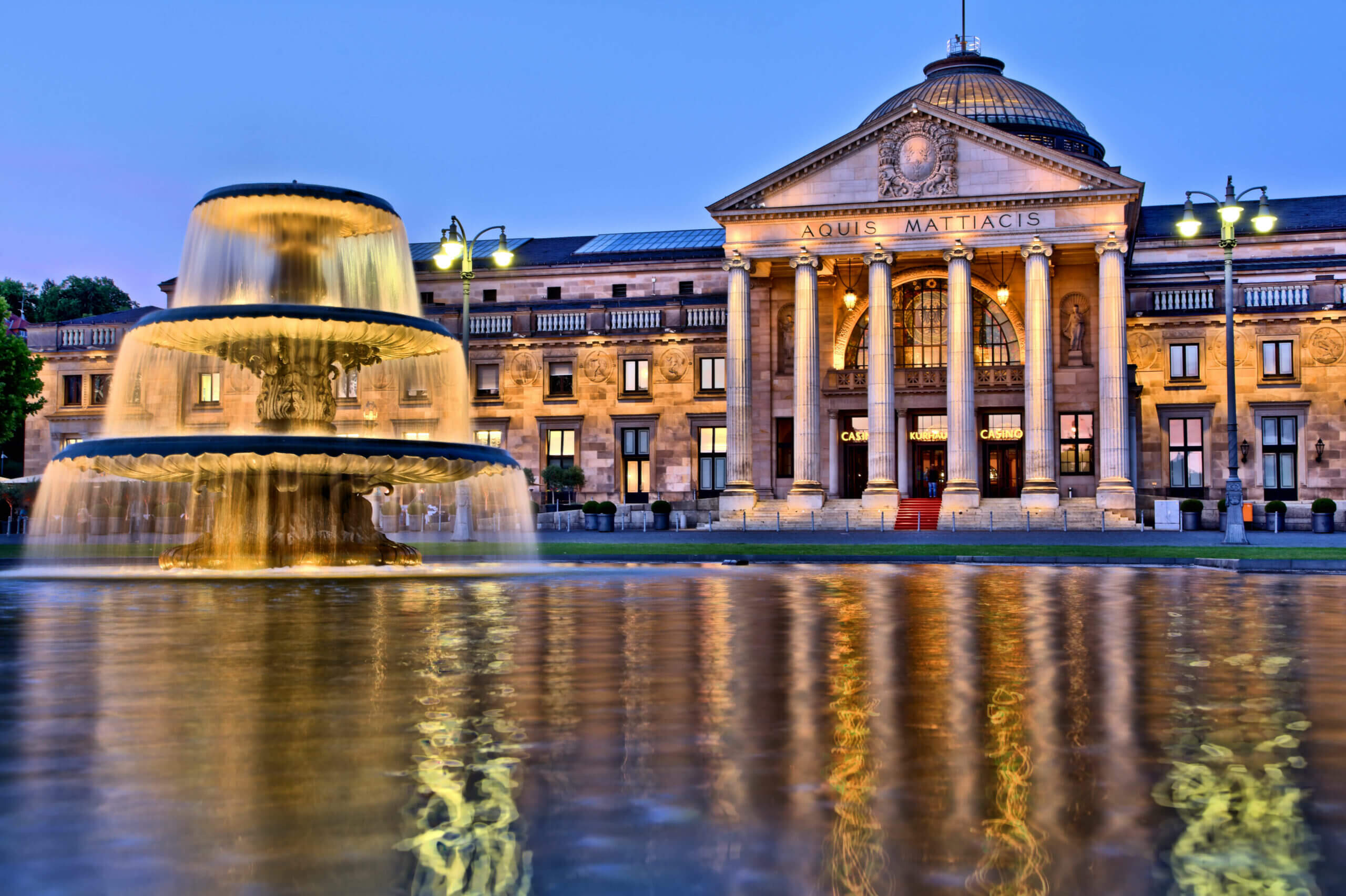 Escort Wiesbaden - Beleuchtetes Wiesbadener Kurhaus in den Abendstunden mit Brunnen