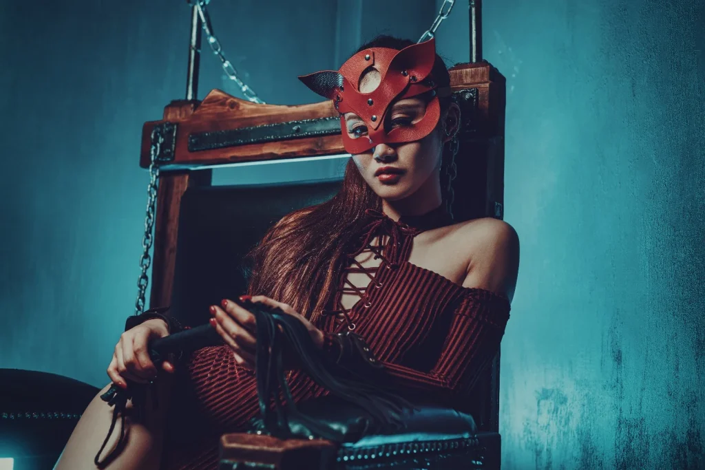 Eine Dame, verhüllt mit einer roten Ledermaske, schaut selbstbewusst in die Kamera und sitzt auf einem Holzsessel. Die Szene ist in schummriges, blaues Licht gehüllt – ein Beispielbild für BDSM-Escort.