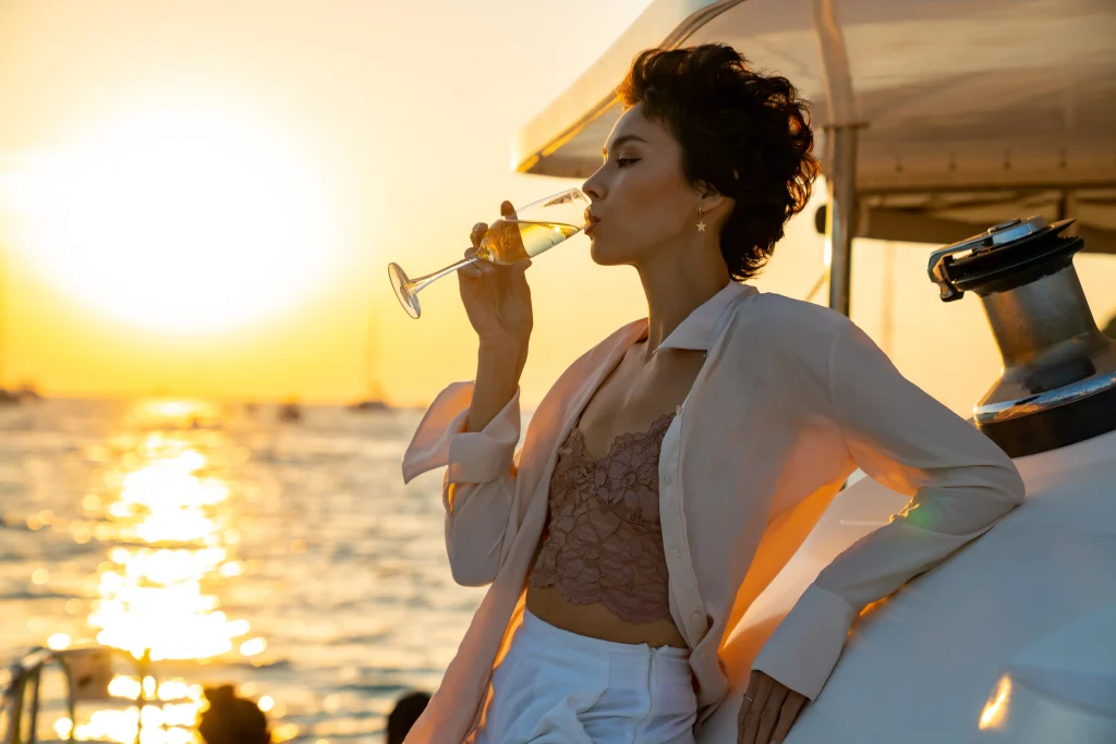 Eine Dame mit kurzen braunen Haaren trinkt genüsslich einen Cocktail in der abendlich untergehenden Sonne. Dabei trägt sie eine weiße Hose und eine leichte Sommerbluse. Ein Beispielbild für Escort-Reisebegleitung.