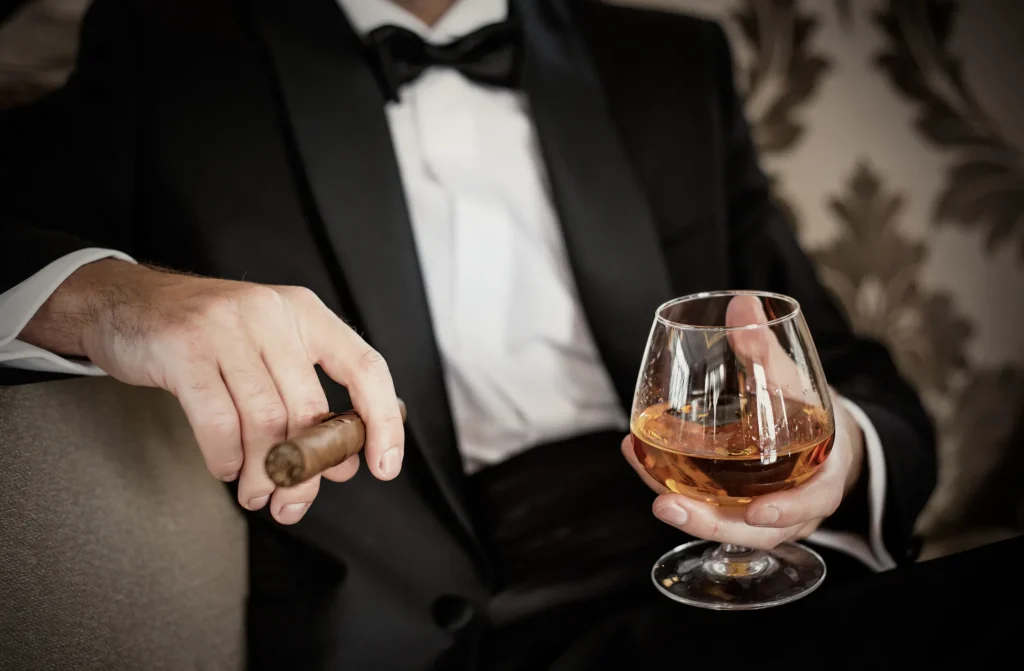 Ein Mann im Smoking hält ein Glas Whiskey und eine Zigarre in der Hand, lässig und elegant. Ein Beispielbild für Escort Stuttgart.