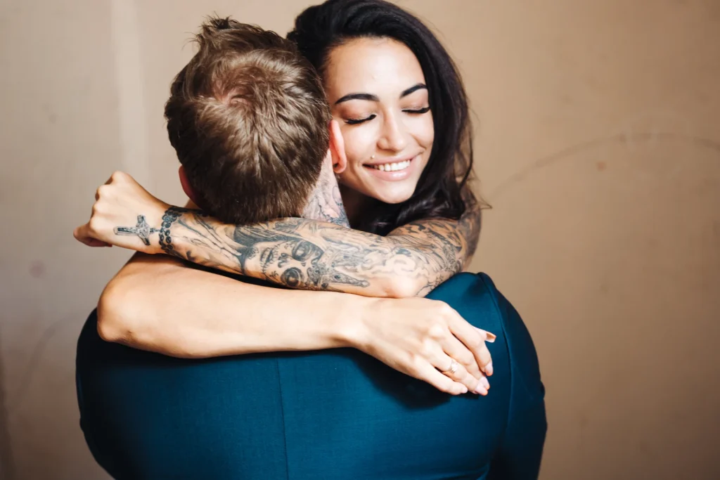 Tätowierte Frau umarmt liebevoll einen Mann in blauem Anzug und lächelt mit geschlossenen Augen – Symbolbild für Nähe und Zärtlichkeit im Rahmen der girlfriend experience (GFE).